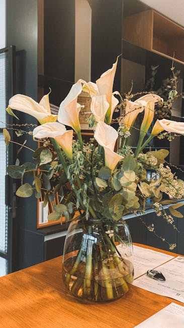 A large, elegant floral arrangement of cream-colored Calla lilies with smooth, trumpet-shaped petals and long, slender green stems, complemented by sprigs of small white filler flowers and round, silv
