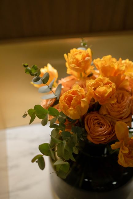 A bouquet of fresh, vibrant orange ranunculus flowers arranged in a dark glass vase, complemented by sprigs of eucalyptus with round silvery-green leaves. The flowers are densely packed, showcasing th