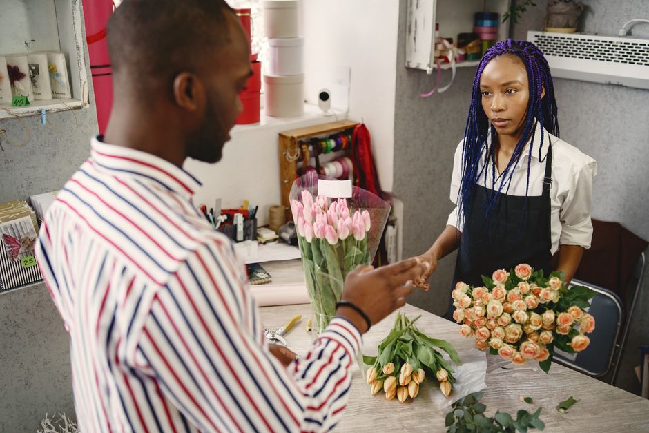 A man in a striped shirt interacts with a female florist inside a floral shop, exchanging a thank you or payment gesture. The table in front of them displays a bouquet of pink tulips wrapped in clear 