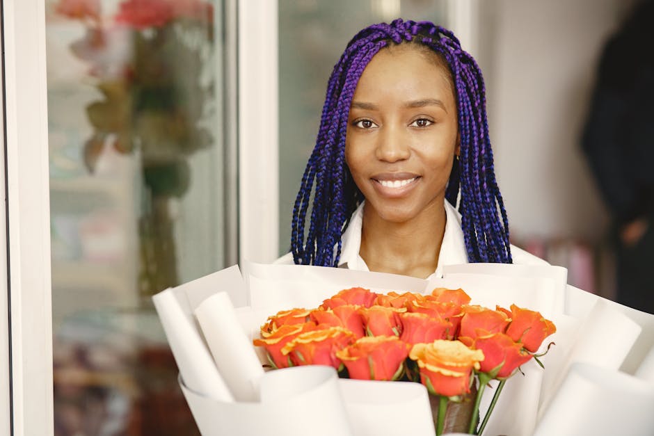 A young woman with medium-dark skin and vibrant purple and black braided hair is smiling warmly while holding a large bouquet of fresh orange and yellow roses wrapped in white paper. She appears to be
