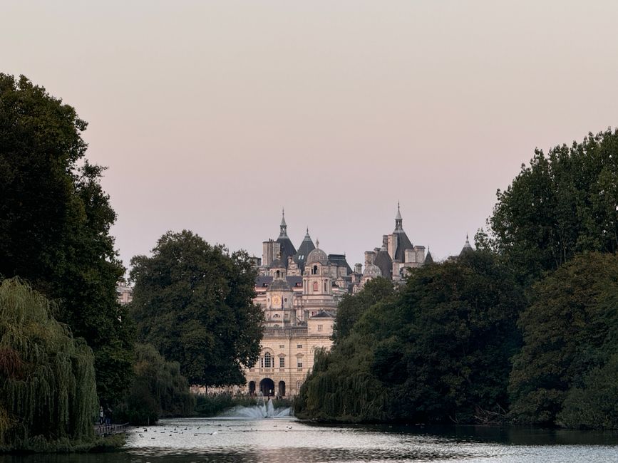 A scenic view of a historic castle with multiple turrets and spires, set against a pastel-colored sky during dawn or dusk, surrounded by lush green trees. In the foreground, a calm river reflects the 